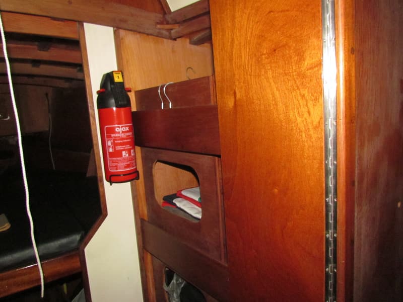 Een rode Ajax brandblusser hangt aan de wand in een houten interieur, met een witte lijn links en opgevouwen kleding in een…