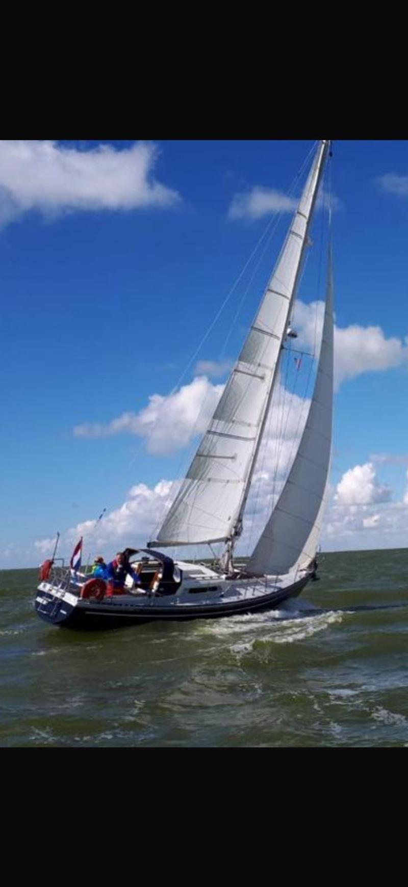 Een zeilboot met twee zeilen en een Nederlandse vlag vaart op een water met golven onder een blauwe lucht met wolken.