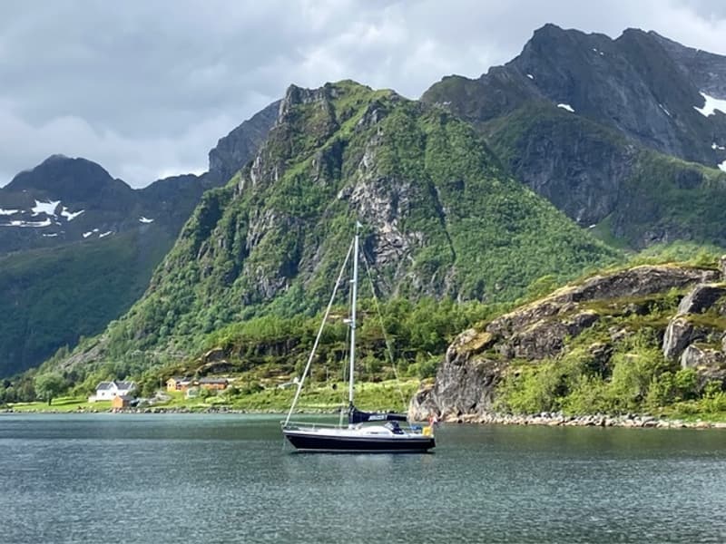 Een zeilboot met donkere romp en witte opbouw ligt in het water, omringd door bergen met groene begroeiing.