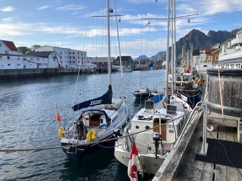 Twee zeilboten en meerdere vissersboten liggen afgemeerd aan een houten steiger in een haven, met bergen op de achtergrond.