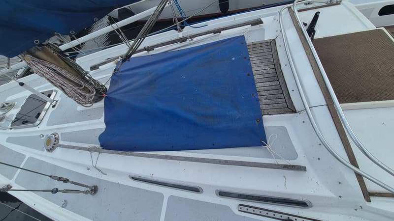 Een bovenaanzicht van een wit zeiljacht met een blauw zeil en een blauw dekzeil, met touwen en houten details.