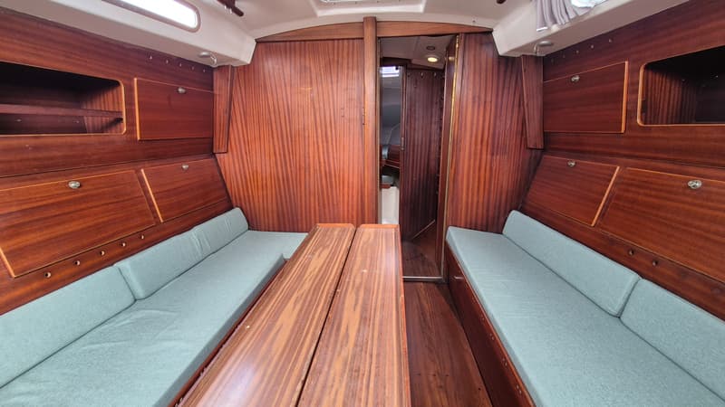 De kajuit toont twee banken met lichtblauwe kussens, een houten tafel en houten wanden met opbergvakken. — Contest 36 Ketch