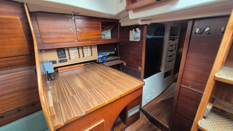 De foto toont een houten interieur van een boot met een tafel, schakelpaneel, opbergruimte en een koelkast.