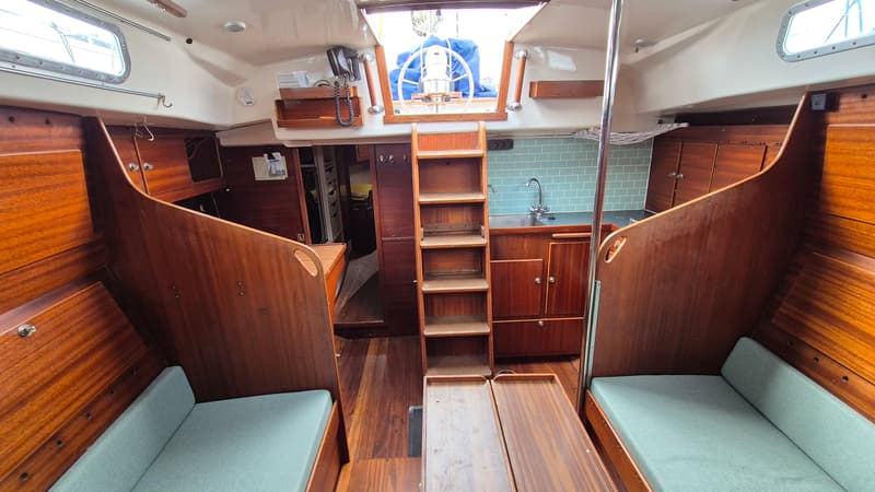 De foto toont het interieur van een boot met houten wanden, vloer en meubilair, en twee banken met lichtgroene kussens.