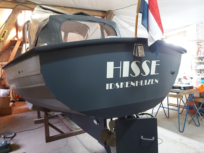 Een donkergrijze boot met de naam "HISSE IDSKENHUIZEN" op de spiegel, een Nederlandse vlag en een schroef is zichtbaar.