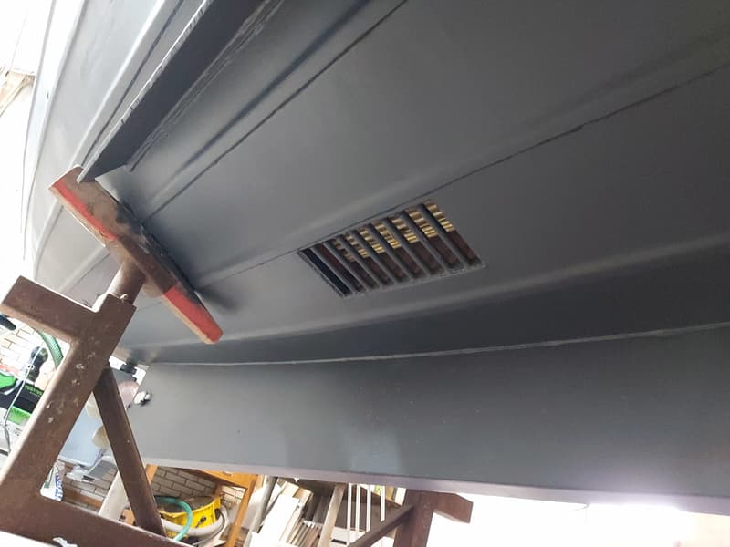 De onderkant van een grijze boot met een rechthoekig rooster is zichtbaar, ondersteund door een roestbruine bok.