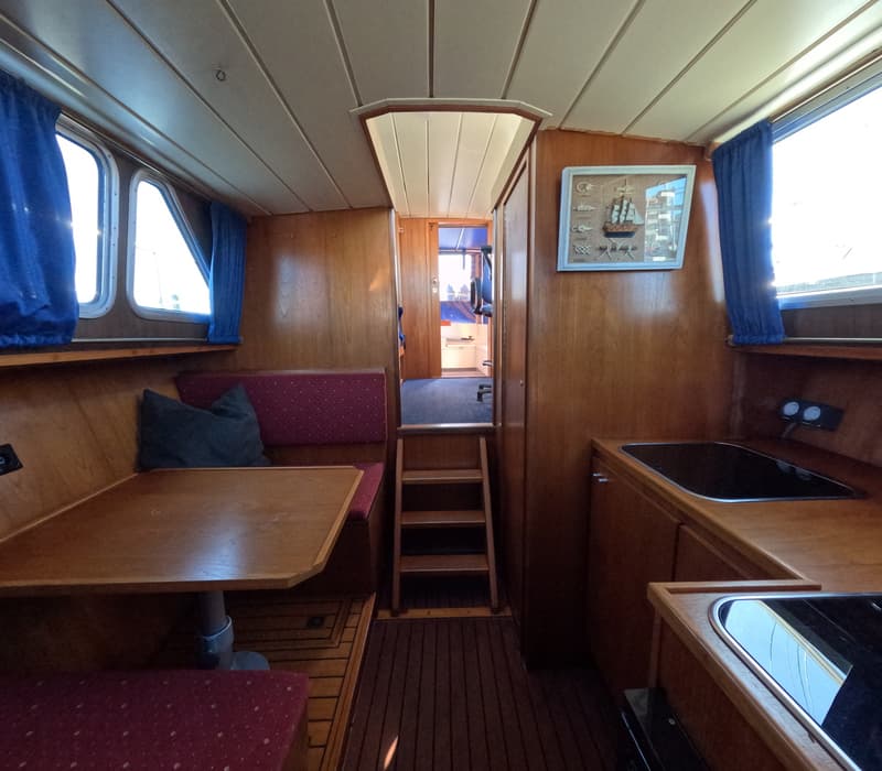 De foto toont het interieur van een boot met houten wanden, een zithoek, een kombuis en een trap naar een ander compartiment.