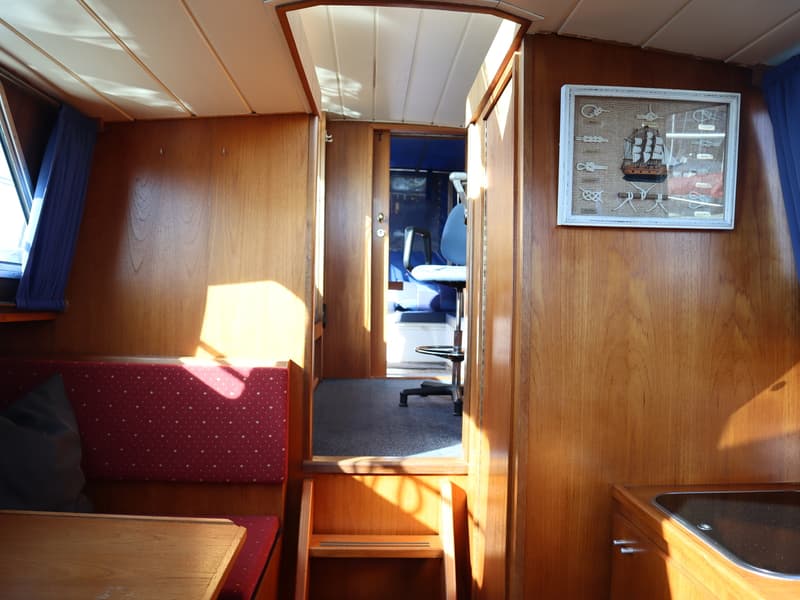 De foto toont het interieur van een boot met houten wanden, een rode bank, een tafel en een doorkijk naar een andere ruimte.