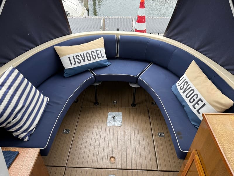 Een bootinterieur met een U-vormige zithoek met donkerblauwe kussens, twee kussens met de tekst "IJSVOGEL" en een gestreept…