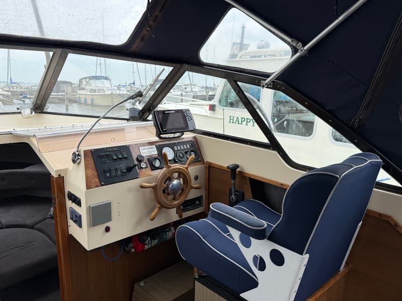 De foto toont de stuurstand van een boot met een houten stuurwiel, diverse meters en schakelaars, en een blauwe stoel.