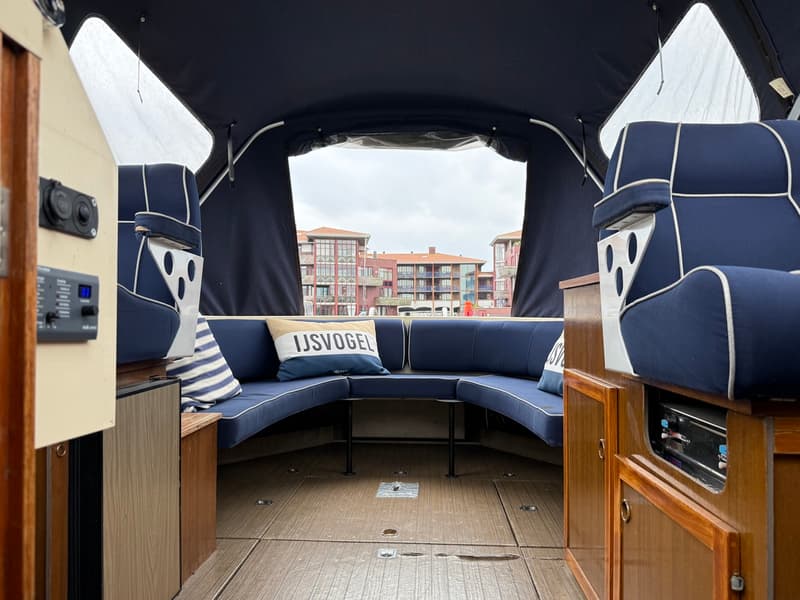 De foto toont het interieur van een boot met blauwe kussens, een kussen met "IJSVOGEL" erop, en houten details. — Joda 7500