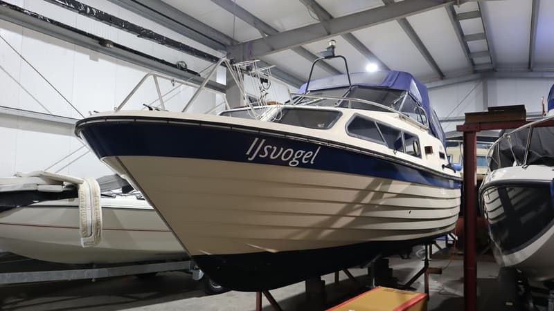 Een beige boot met een blauwe streep en de naam "IJsvogel" staat op bokken in een loods. — Joda 7500