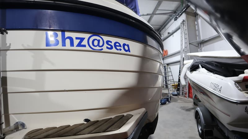 Een witte boot met blauwe accenten en de tekst "BhZ@sea" is zichtbaar, met een houten instap en een andere boot op de…