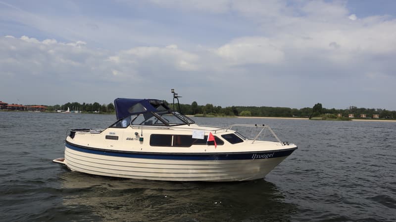 Een crèmekleurige motorboot met blauwe accenten en een blauwe kap vaart op een waterlichaam onder een bewolkte lucht.