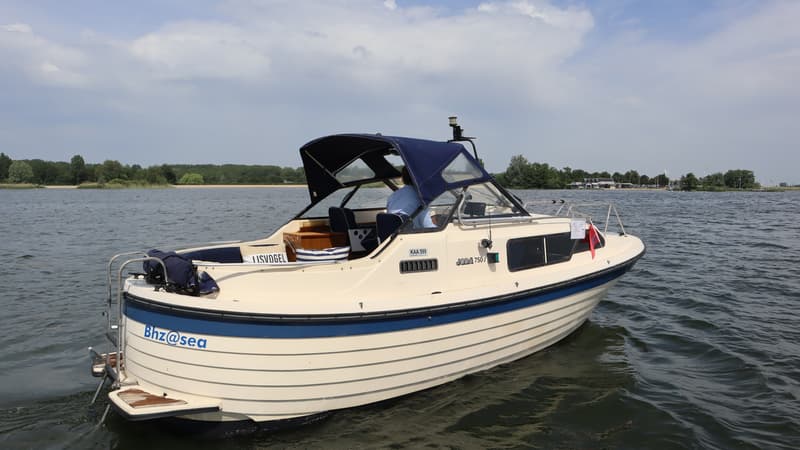Een beige motorboot met een blauwe kap en een persoon aan het roer vaart op rustig water onder een bewolkte hemel.