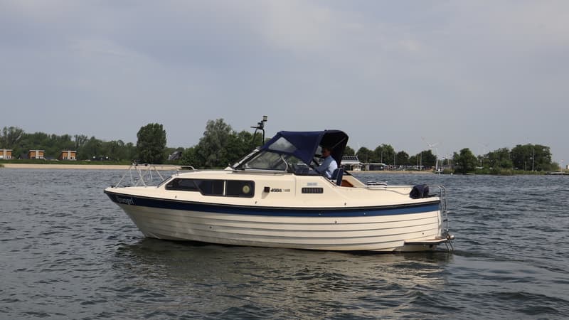 Een beige motorboot met blauwe accenten en een blauwe kap vaart op het water met een persoon aan het roer. — Joda 7500