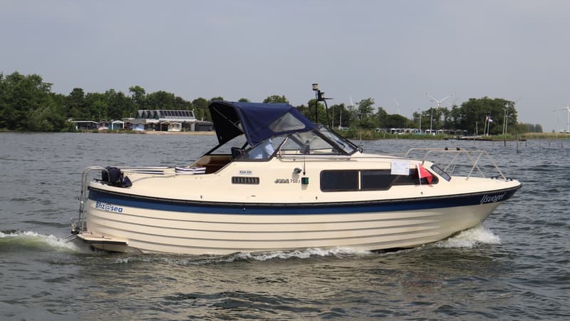 Een beige motorboot met blauwe accenten en een blauwe kap vaart op het water, met een kustlijn op de achtergrond. — Joda 7500