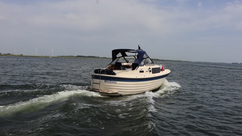 Een crèmekleurige motorboot met blauwe accenten en een blauwe kap vaart over water, met windmolens op de achtergrond.