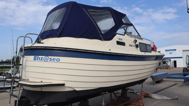 Een crèmekleurige boot met een blauwe streep en een blauwe kap staat op een trailer, met de naam "BhZ@sea" op de… — Joda 7500