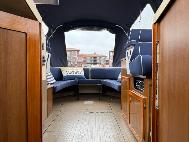 De foto toont het interieur van een boot met een blauwe bank, een kussen met "IJSVOGEL" erop, en houten panelen. — Joda 7500
