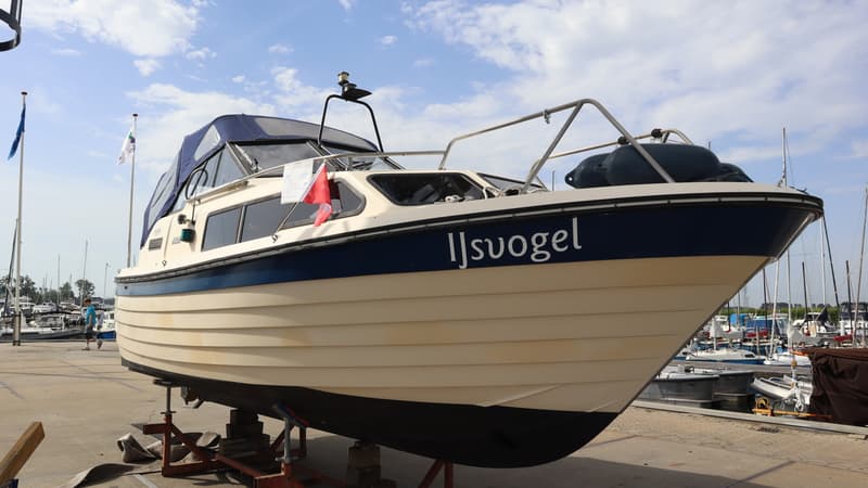 Een crèmekleurige boot met een blauwe streep en de naam "Ijsvogel" staat op bokken op de wal. — Joda 7500