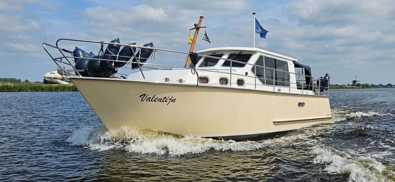Een beige boot met de naam "Valentijn" vaart over water, met een molen op de achtergrond. — Bach 1050 OK