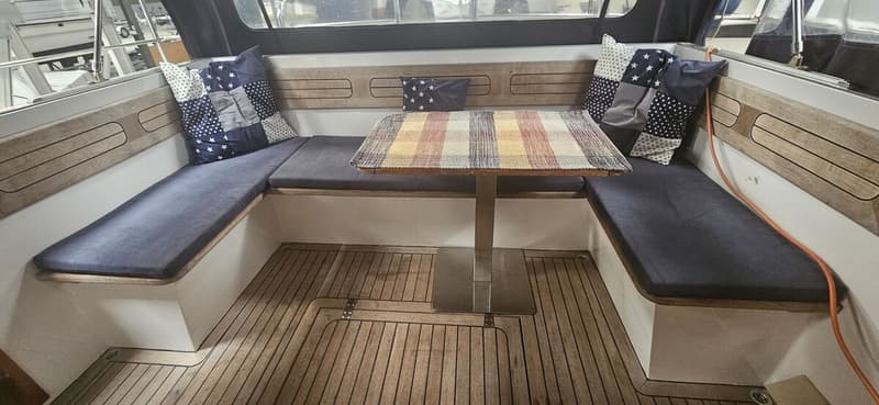 Een interieur van een boot met een U-vormige zitbank met donkerblauwe kussens, een tafel en een houten vloer. — Bach 1050 OK