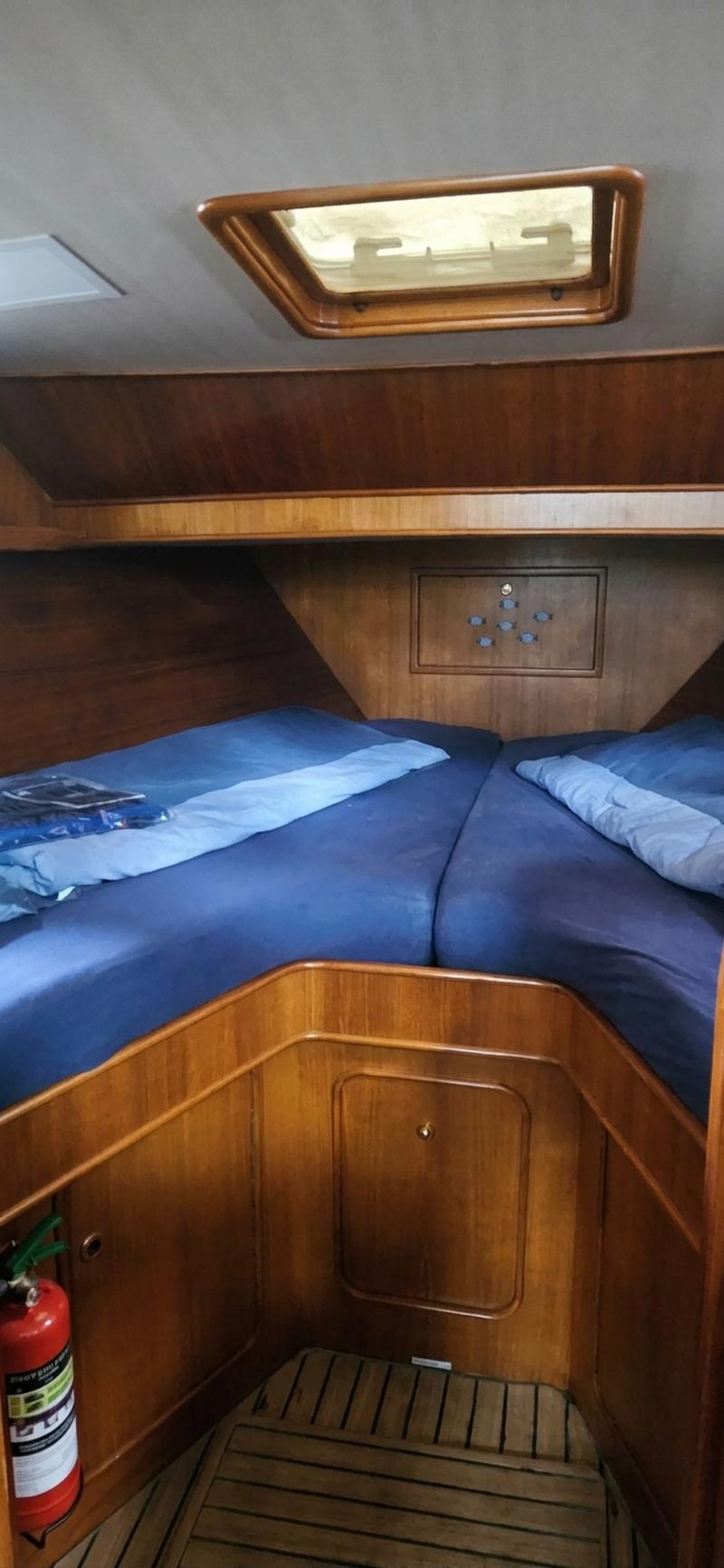 De foto toont een kajuit met twee bedden met blauwe hoezen, een houten interieur, een dakluik en een brandblusser.