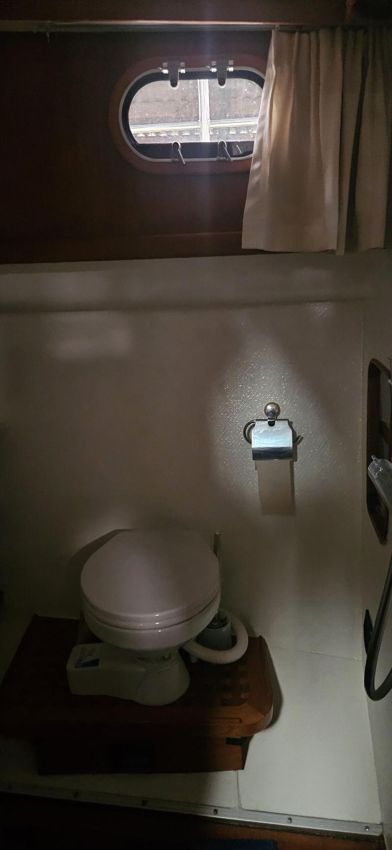 Een boottoilet met een toiletrolhouder en een patrijspoort met een gordijn erboven.
