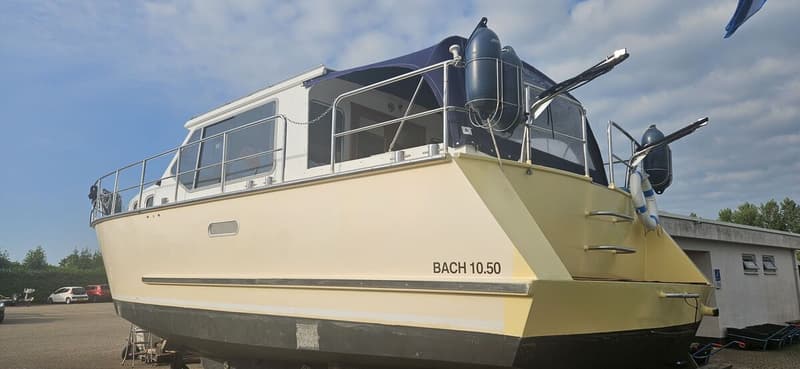 Een beige boot met de tekst "BACH 10.50" op de achtersteven, staat op het droge onder een blauwe lucht. — Bach 1050 OK