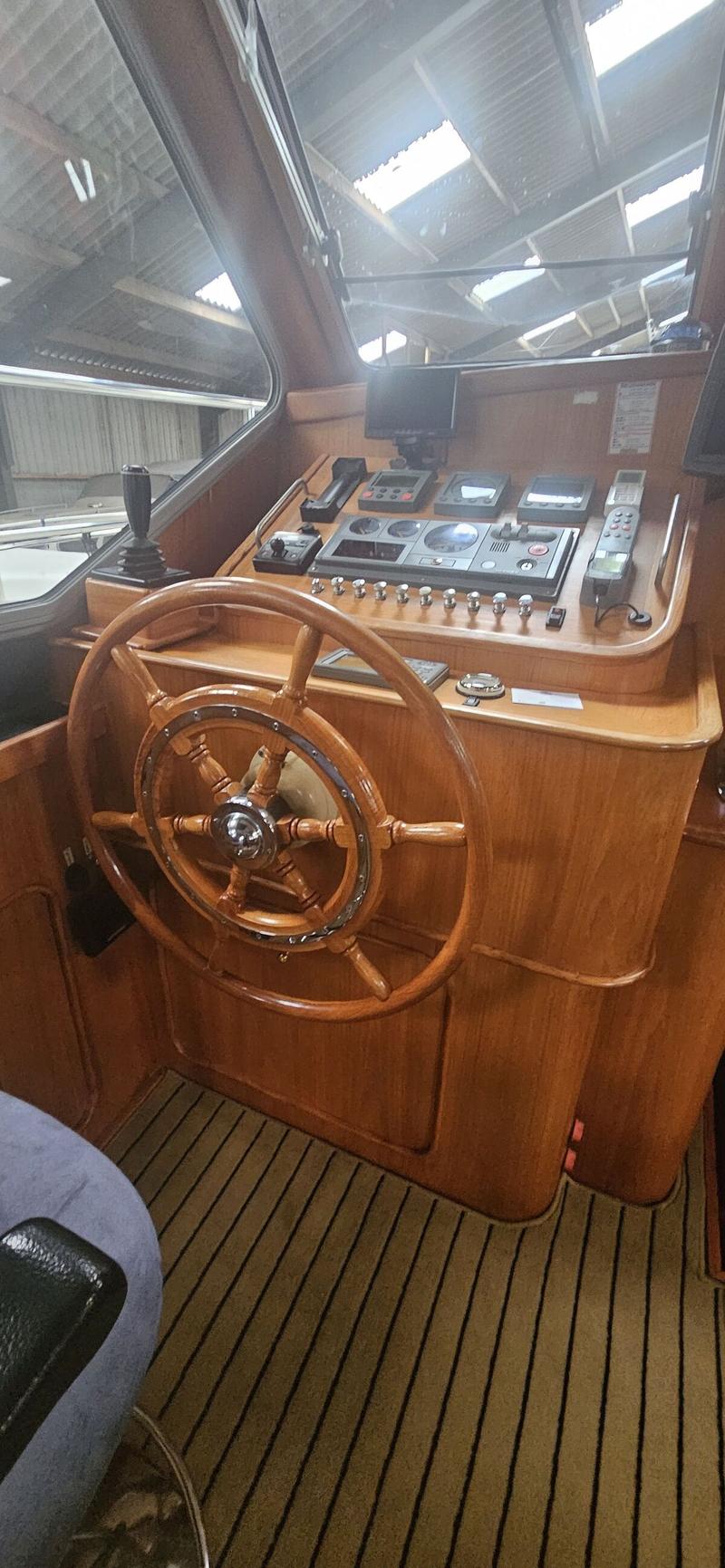 Een houten stuurwiel en dashboard met diverse meters en knoppen, met een gestreepte vloer en een raam dat een dak van een…