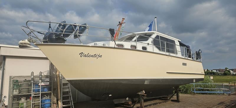 Een crèmekleurige motorboot met de naam "Valentijn" ligt op de wal, ondersteund door stutten. — Bach 1050 OK