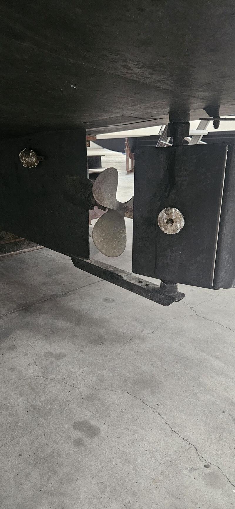 Een detailfoto van de onderkant van een boot met een tweebladige schroef en twee roeren, boven een betonnen vloer.