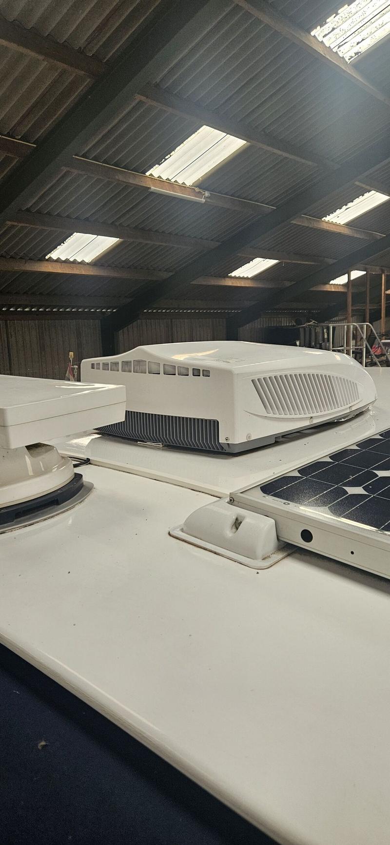 Een wit dak van een boot met een airconditioningunit, een zonnepaneel en een antenne, onder een golfplaten dak.