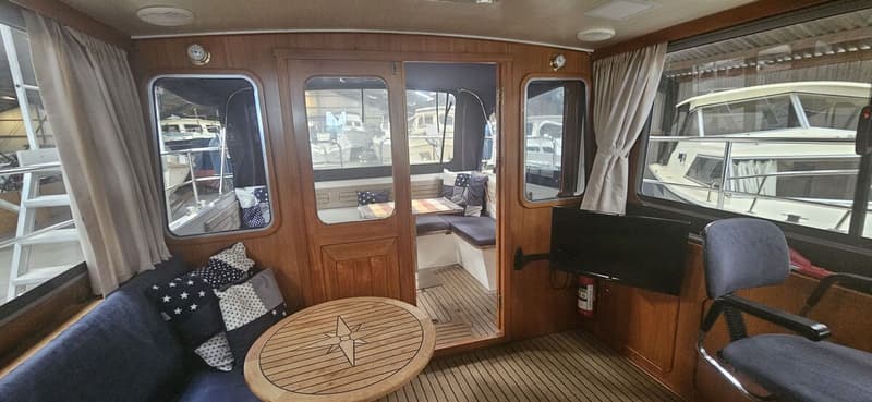De foto toont het interieur van een boot met houten wanden, ramen, een zithoek, een tafel en een stuurstoel. — Bach 1050 OK