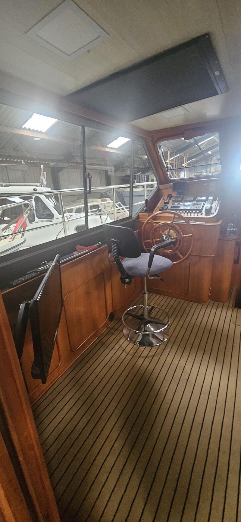 De foto toont het interieur van een stuurhut met een houten stuurwiel, een draaibare stoel en een houten vloer met zwarte…