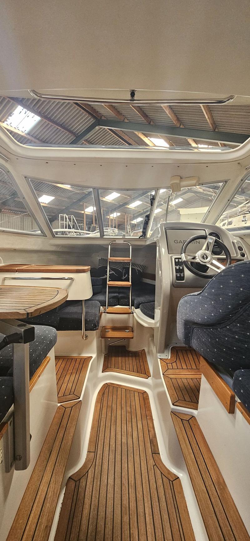 De foto toont het interieur van een boot met een stuurwiel, een Garmin-scherm, houten vloeren en blauwe kussens.