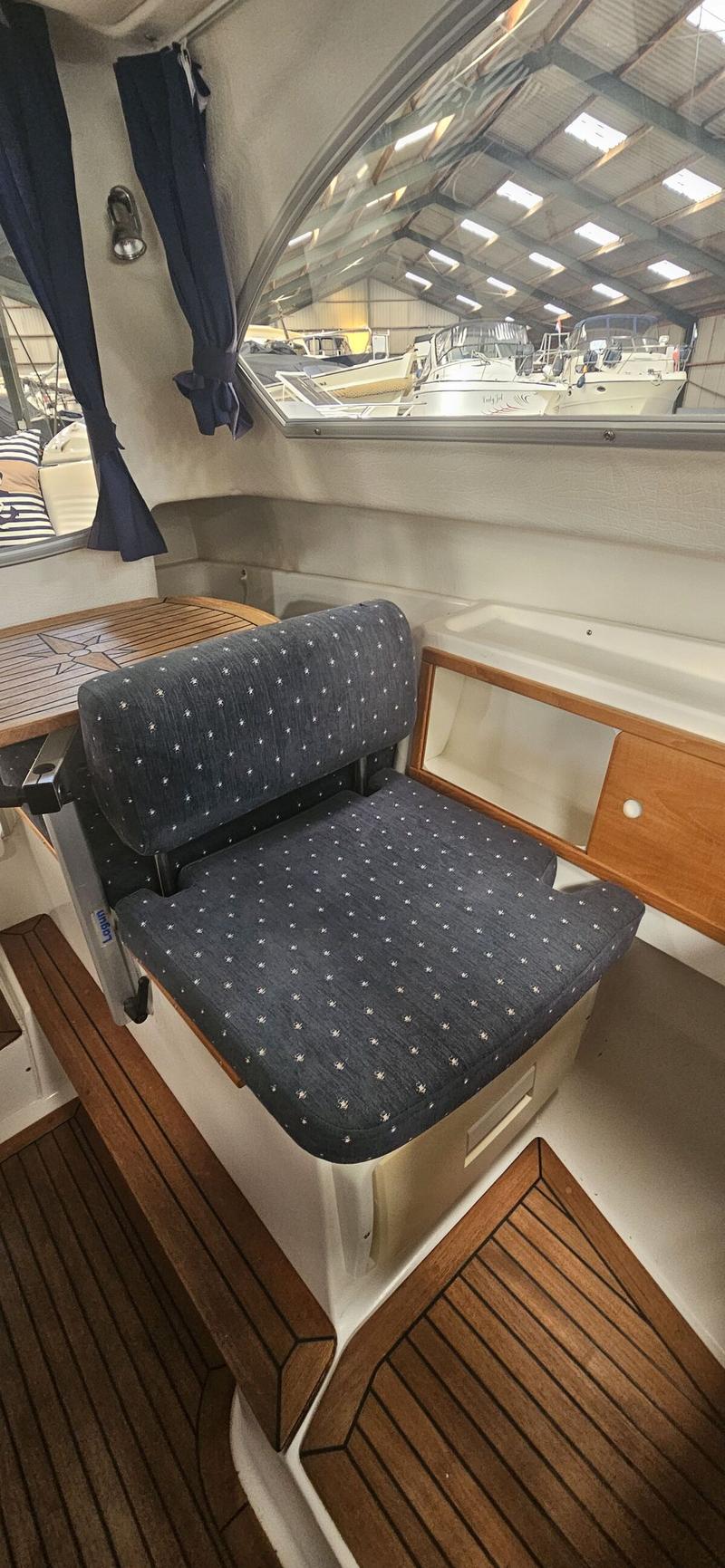 Een interieur van een boot met een donkerblauwe gestoffeerde stoel, een houten tafel en een raam met uitzicht op een loods.