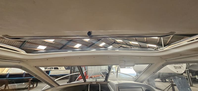 De foto toont het interieur van een boot met een open dak en een voorruit, met uitzicht op een loods met andere boten.