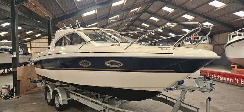 Een witte boot met een donkerblauwe streep en gouden accenten staat op een trailer in een loods. — Yamarin 68C
