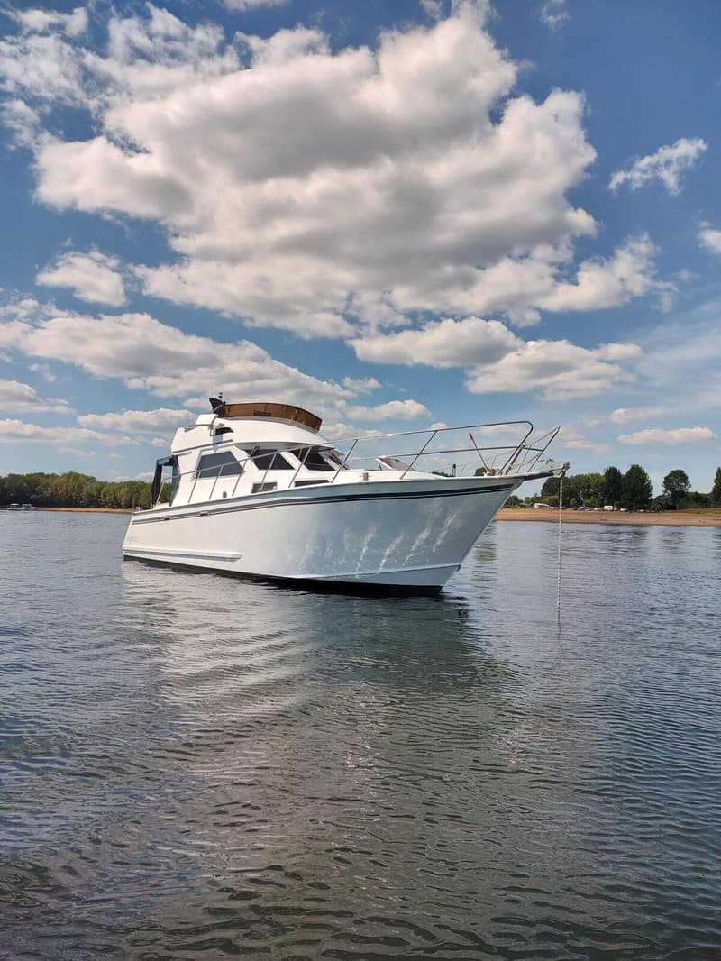 Een witte motorboot ligt op het water onder een blauwe lucht met witte wolken, met een groene oever op de achtergrond.