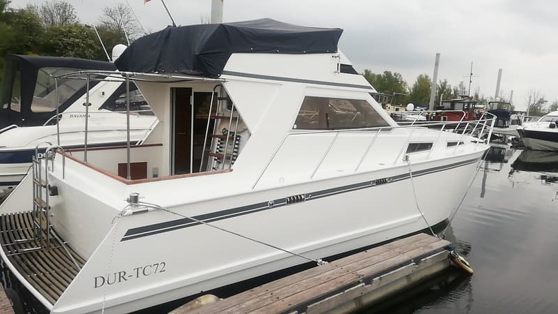 Een witte motorboot met een zwarte kap ligt aangemeerd aan een houten steiger, met andere boten op de achtergrond.