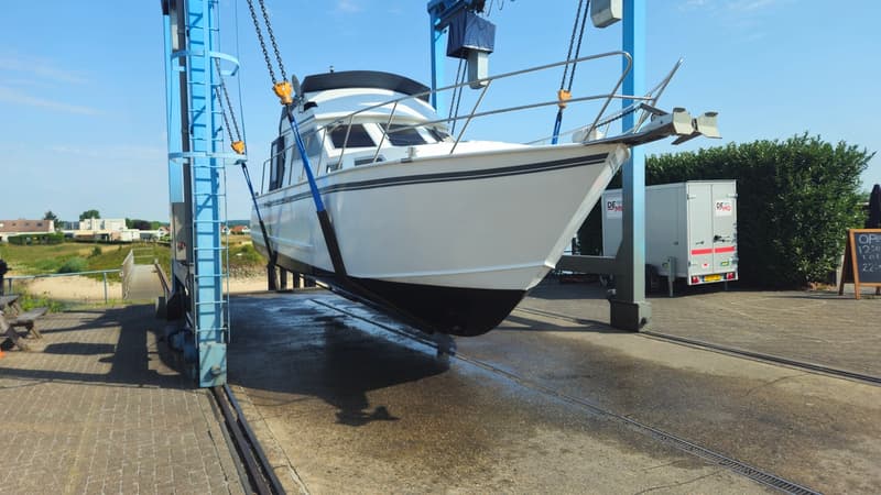 Een witte motorboot met zwarte strepen en een zwarte onderkant hangt in een blauwe botenlift boven een natte betonnen…
