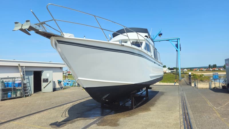 Een witte boot met zwarte strepen en een zwart onderwaterschip staat op een trailer op de wal, naast een gebouw en een…