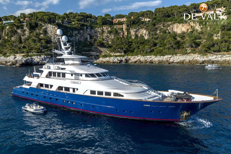 Een grote blauw-witte motorjacht met de naam "L'albatros" ligt in het water voor een rotsachtige kustlijn met bebossing.