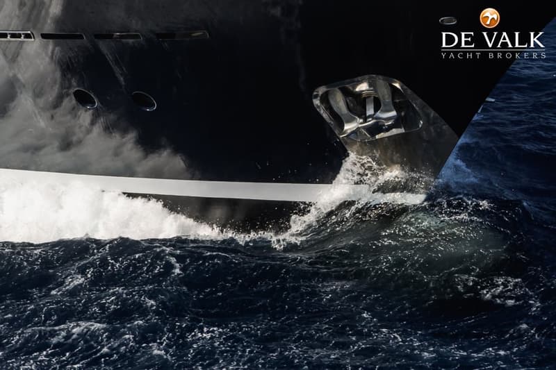 Een donkere boot met een anker in de boeg vaart door woelig water, met het logo "DE VALK YACHT BROKERS" rechtsboven.