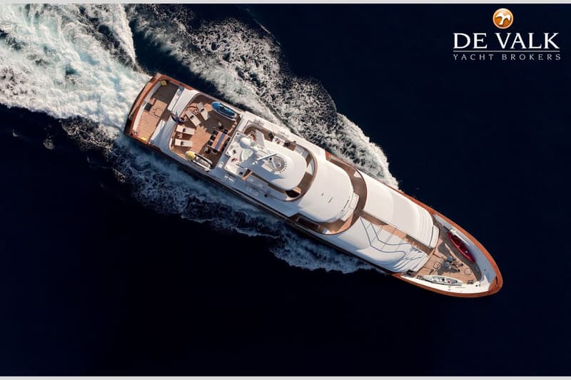 Een luchtfoto van een groot wit motorjacht met houten dekken dat door donkerblauw water vaart en een kielzog achterlaat.