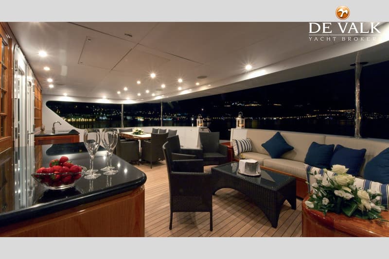 Een luxe jachtdek met zitgedeeltes, een bar met aardbeien en wijnglazen, en uitzicht op een verlichte kustlijn in de nacht.