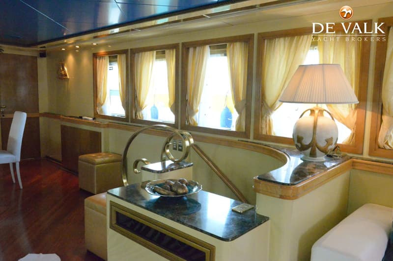 Een interieur van een boot met houten vloer, ramen met gele gordijnen, een lamp en een schaal met decoratieve objecten.