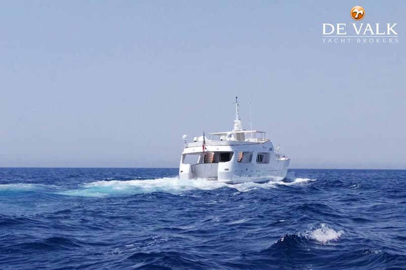 Een witte motorjacht vaart op een blauwe zee, met een vlag aan de achtersteven en een logo van "DE VALK YACHT BROKERS"…